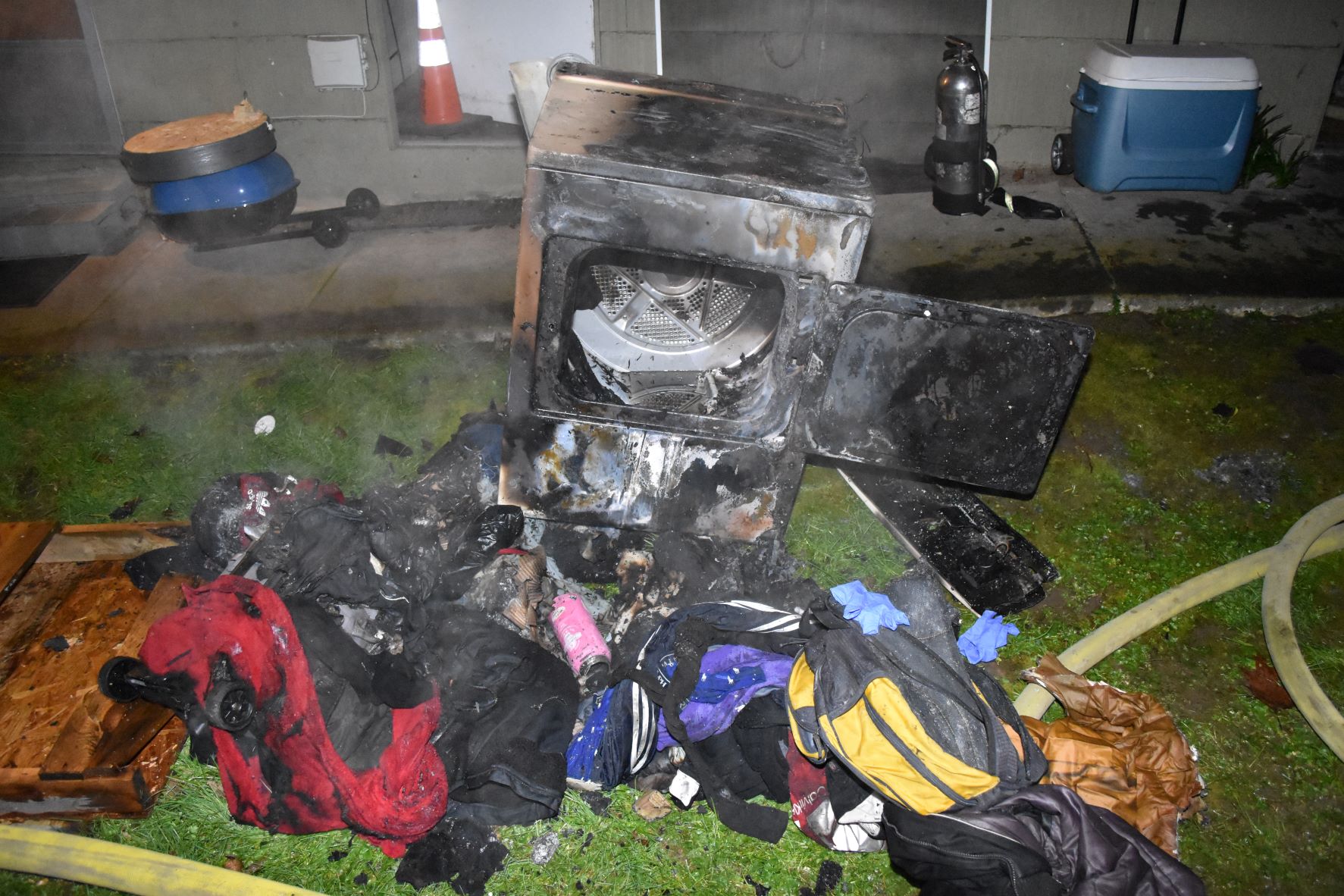 Picture of a dryer that has been extinguished after it was on fire. It sits on a lawn in front of a building with several burnt pieces of clothing. 