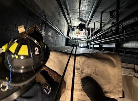 Firefighter looks down elevator shaft as crews work below to rescue occupants from elevator car.
