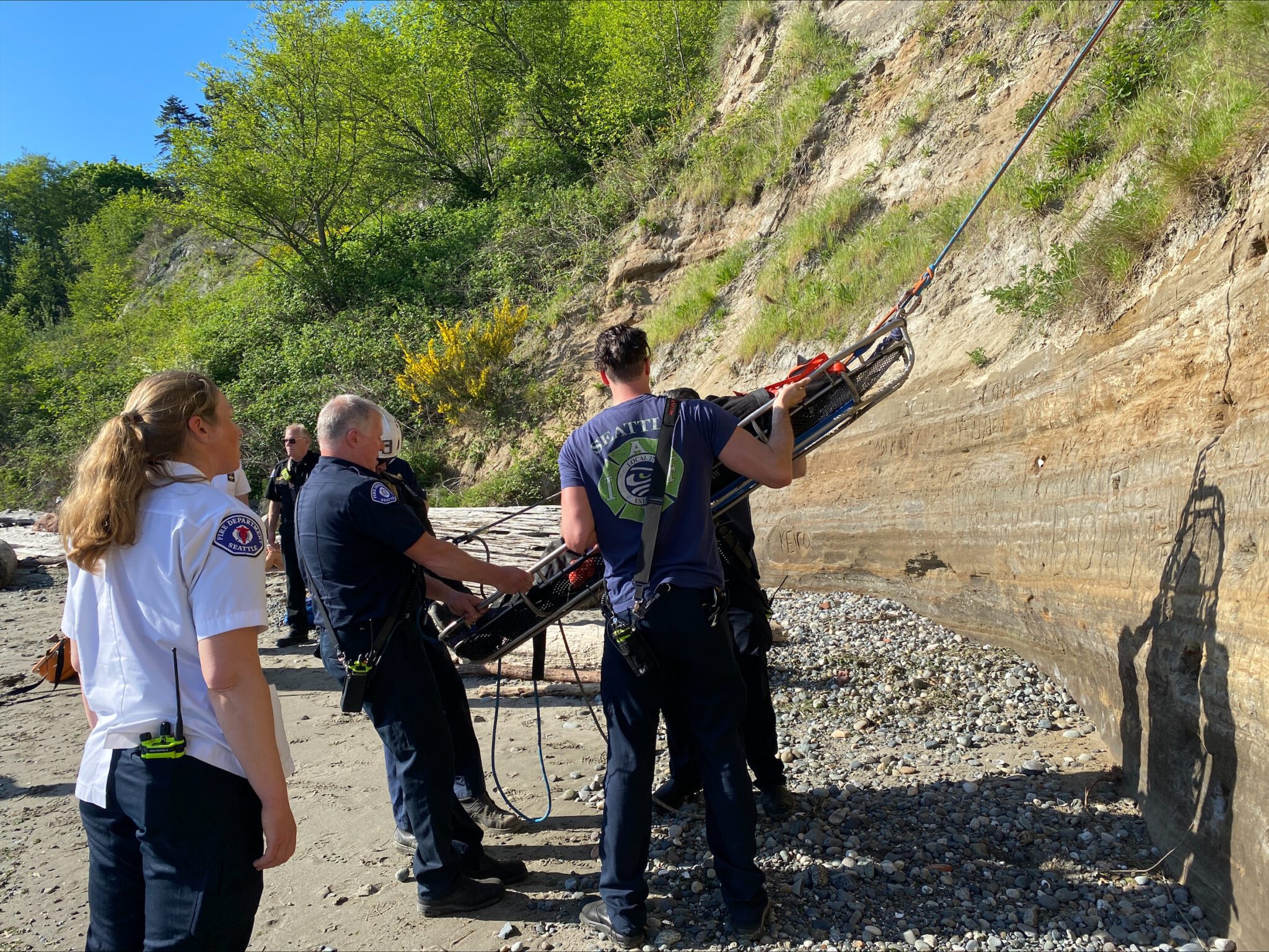 Firefighters hold Stokes basket containing patient being lowered from hillside.