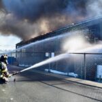 A firefighter sprays water through a fence onto the burning building.