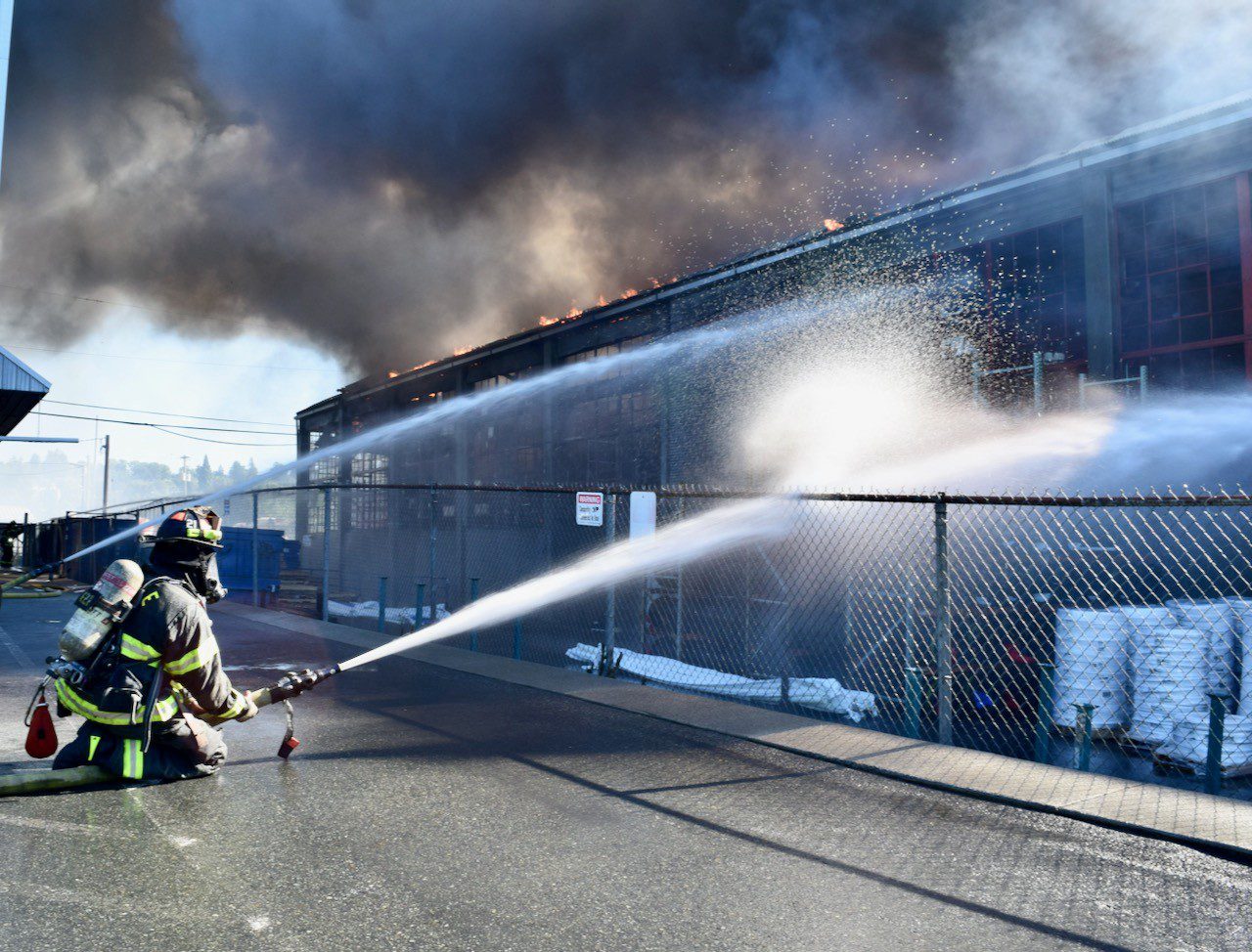 A firefighter sprays water through a fence onto the burning building.