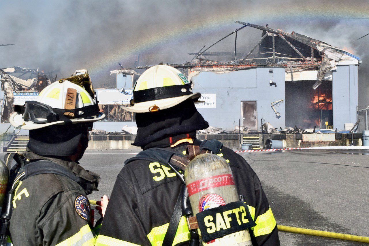Two firefighter stand in front of building on fire as spray from hose line creates a rainbow over them.