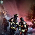 Firefighters talk outside of burning structure. Firefighters in background carry ladder.