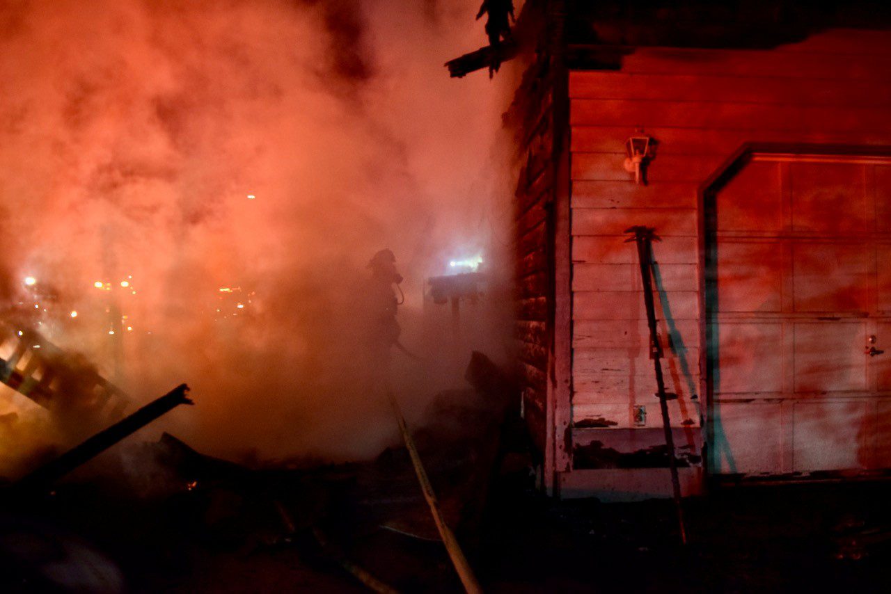 Firefighter with flashlight on exterior of house, surrounded by smoke.